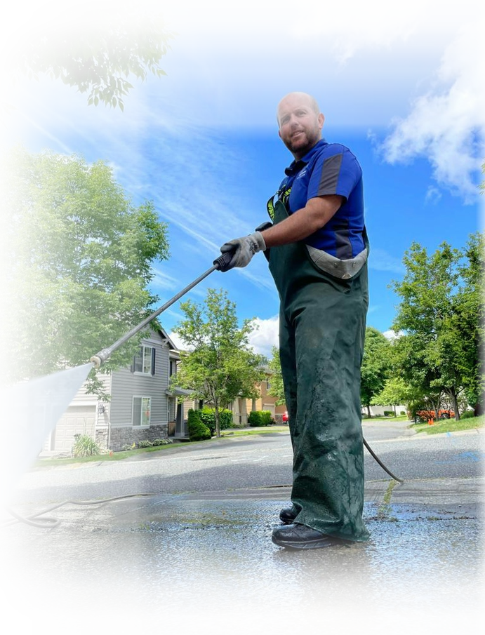 A home exterior cleaning professional uses a water-fed pole to clean tall exterior windows. The man in a blue shirt and patterned shorts is shown in profile, working on large commercial windows.